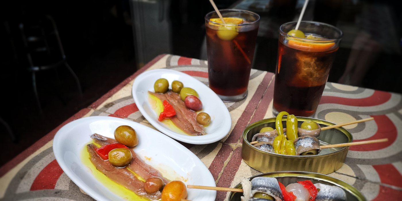 Two glasses of orange-garnished vermouth and tapas, anchovies and olives, on a Barcelona mosaic-tiled table.