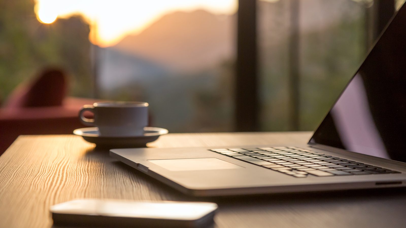 Laptop, coffee cup and phone on a wooden desk before a window at sunset
