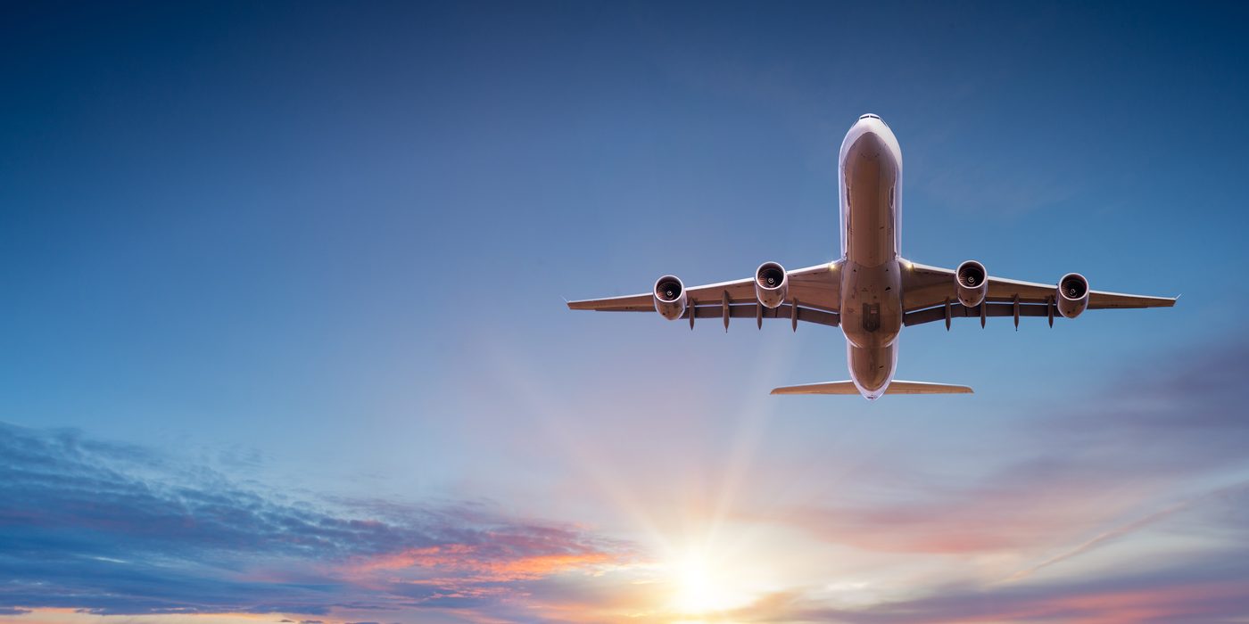 Airliner seen from below against a sunset sky, wings spread, the sun low on the horizon