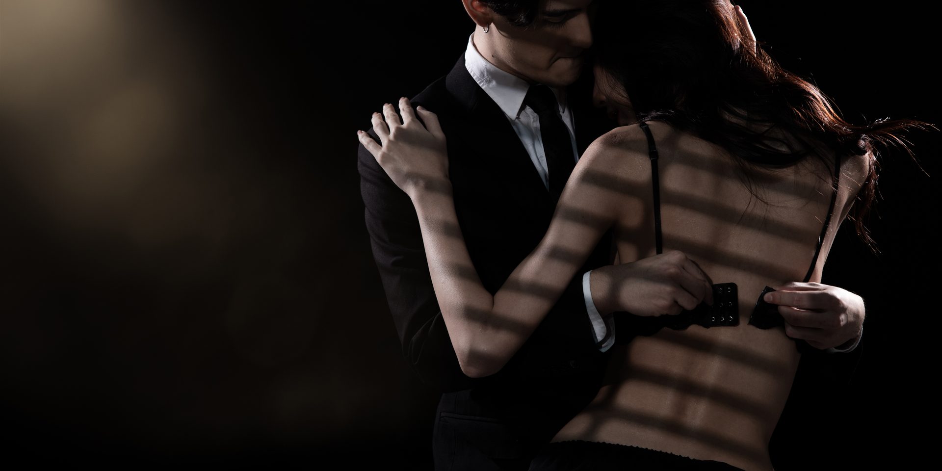 A man in a suit holding his companion close in the striped half-light of a suite.