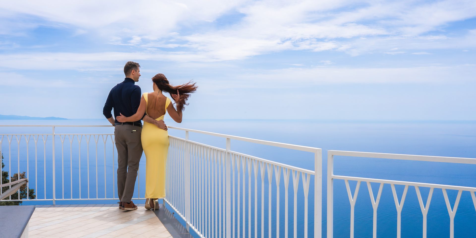 Couple seen from behind on a terrace with white railings high above the sea, she in a long yellow dress, her hair in the wind
