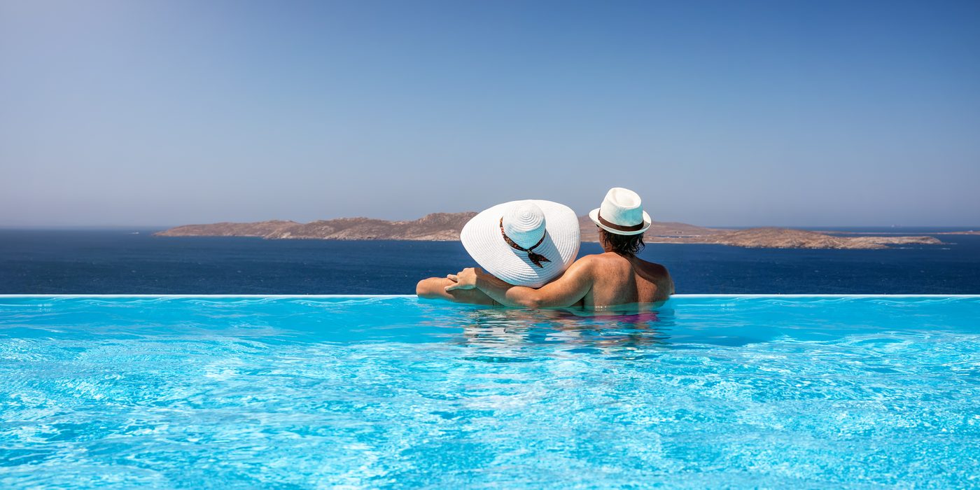 Couple seen from behind at the edge of an infinity pool, in white hats, facing the sea and an island in the distance