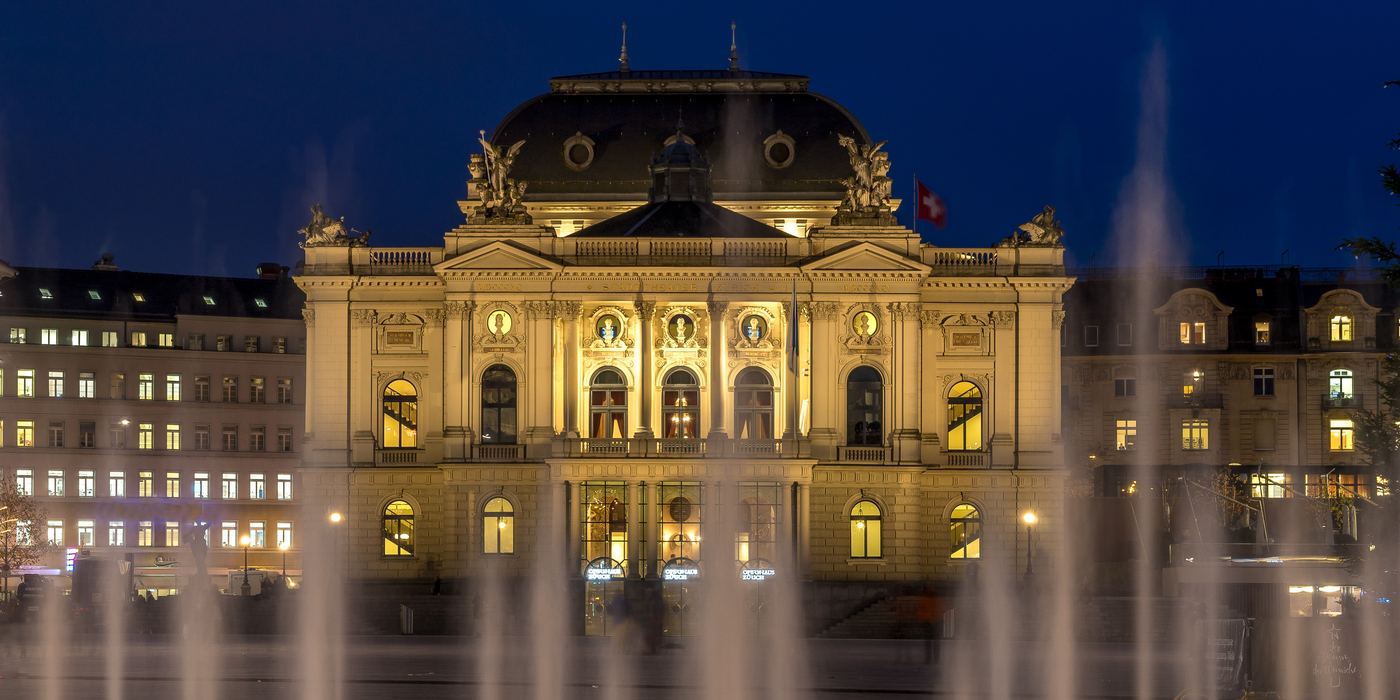 Zurich's Opera House lit up at dusk, the square's fountains in the foreground.
